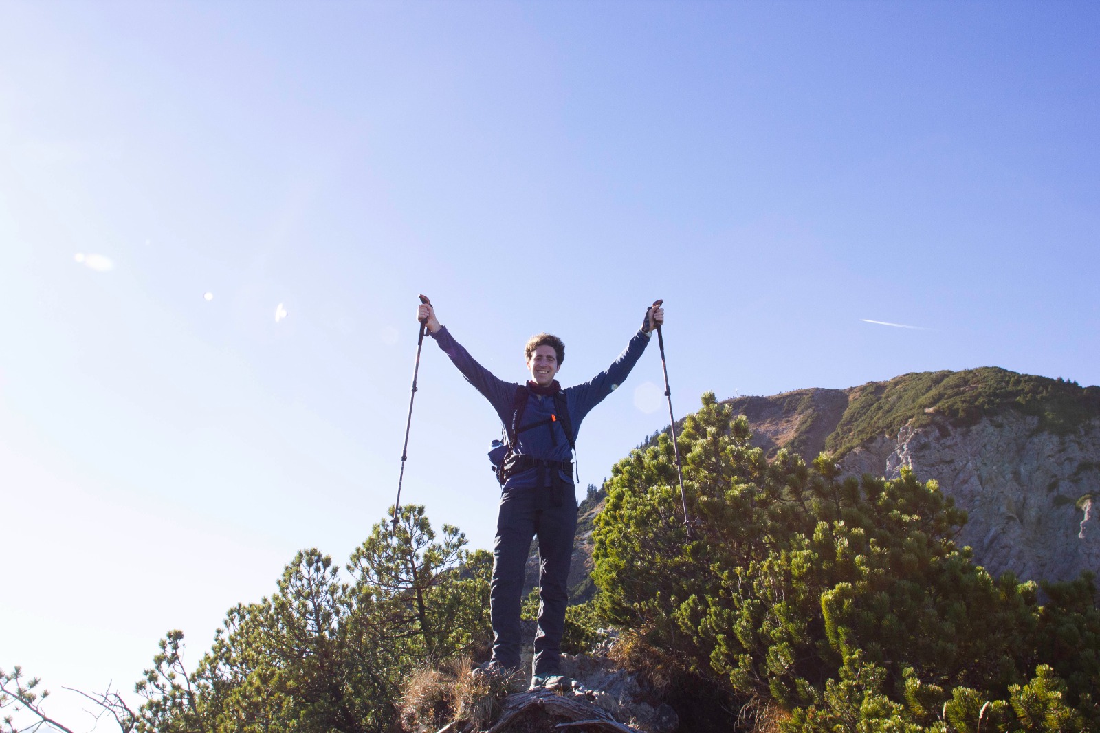 Kevin Mancini standing on a mountain trail with trekking poles raised under a clear blue sky